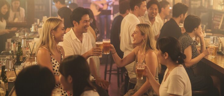 沖縄の北谷アメリカンビレッジ周辺にあるバーで、日本人が外国人客と笑顔で乾杯し、国際交流を楽しんでいる様子。多様な文化が混ざり合う、沖縄ならではの出会いの場を柔らかな光とソフトフォーカスで表現。「沖縄で出会いがない」を解消する多文化交流。