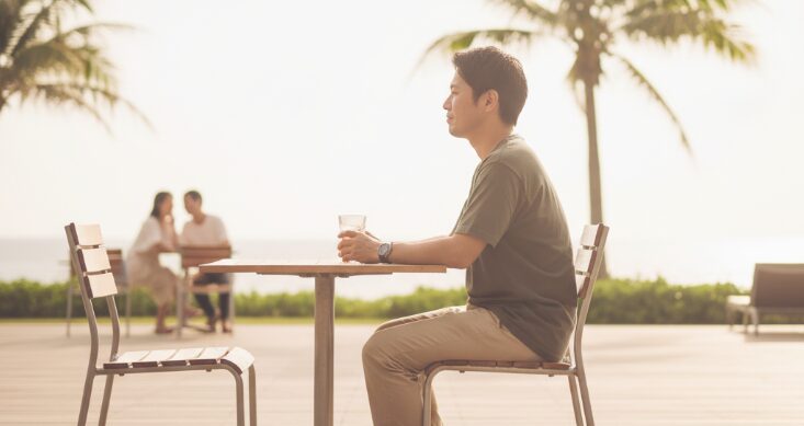 沖縄移住者が直面する出会いの課題を表現する画像。夕暮れ時のビーチで、一人でテーブルに座り物思いにふける男性。遠くにはカップルの姿が見える。