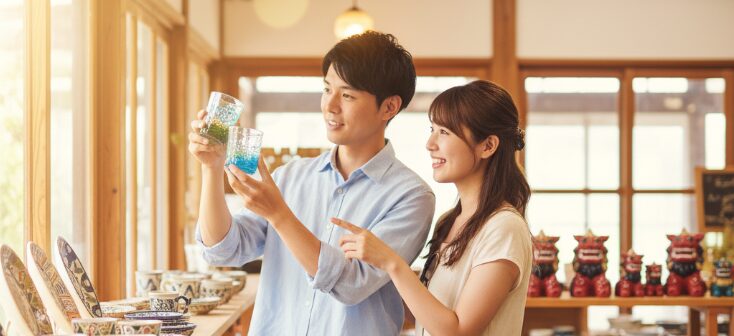 沖縄の地元デートの記念にお土産を選ぶカップル。やちむんや琉球ガラスが並ぶ店内で、楽しそうに品物を選ぶ様子。
