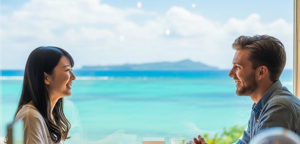 沖縄の絶景を背景に、日本人女性と外国人男性がカフェで楽しそうに会話している様子。沖縄のマッチングアプリを通じて国際的な出会いが生まれる可能性を示唆する、明るく開放的な雰囲気。