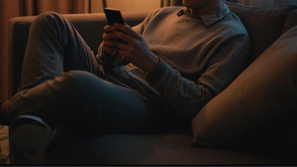 自宅のリビングでスマホを見る男性。マッチングアプリの日記を更新し、多くの足跡がついていることを確認するリラックスした様子。