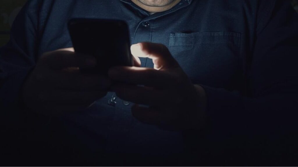 暗い部屋でスマホを見つめ、悩ましげな表情の男性。マッチングアプリのメッセージのコツにおいて避けるべき、相手に負担をかけるNGな状況。