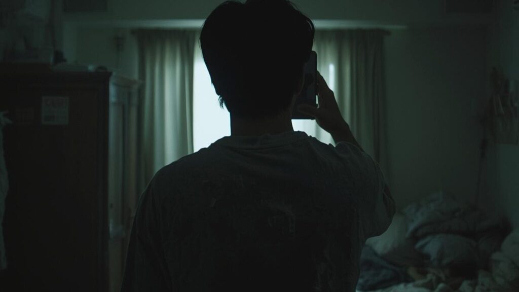 薄暗い部屋で一人、スマートフォンを掲げる日本人男性のシルエット。マッチングアプリの写真で男が避けるべき、孤独感や閉塞感が漂う自撮りの撮影風景。