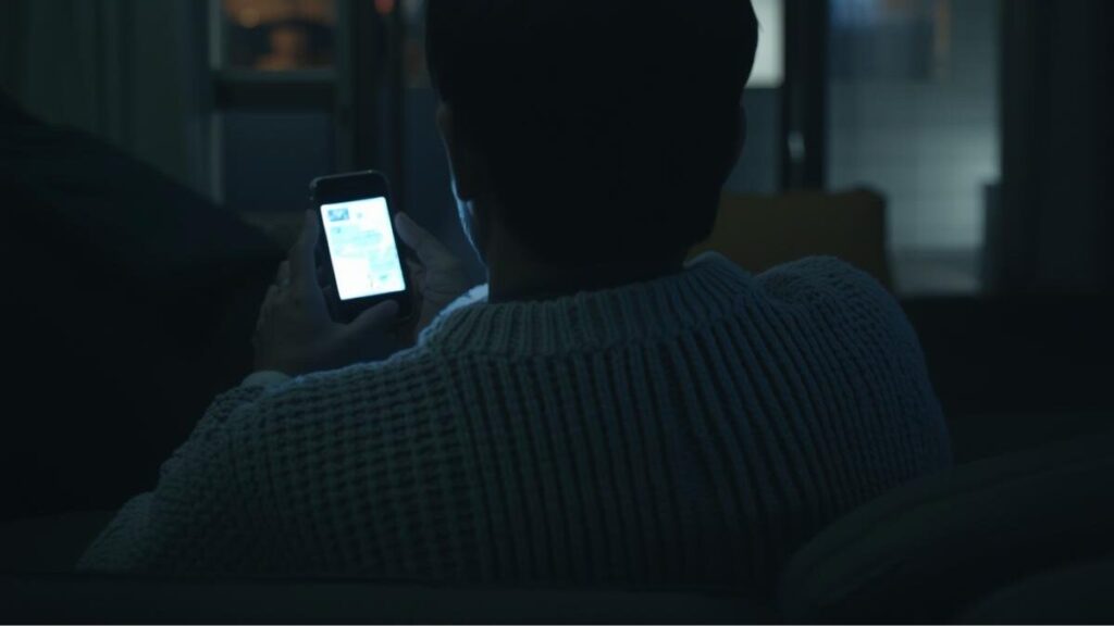 夜のリビングでリラックスしてスマートフォンを見ている日本人男性の後ろ姿。24時間監視体制によって守られ、マッチングアプリの安全性を感じている様子。